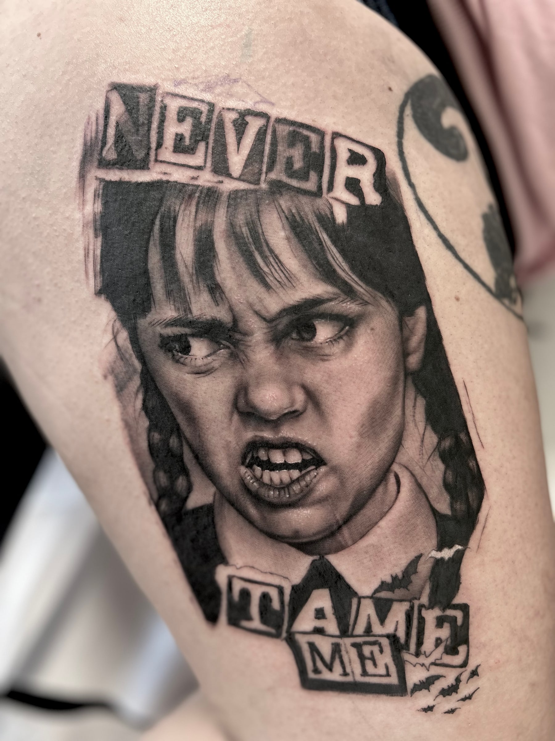 Black and grey portrait of a scowling girl with braids, framed by cut-out letters reading “Never tame me”, on the thigh.